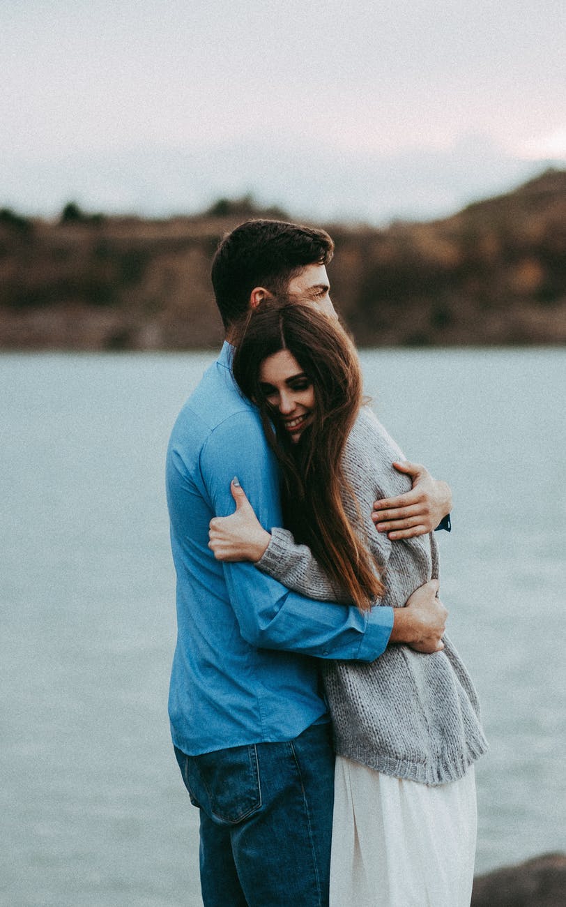 man hugging woman wearing gray sweater near body of water